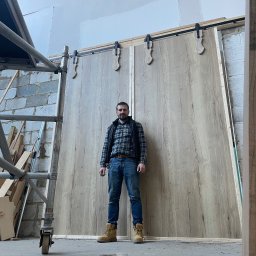 Woodworking - Mężczyzna w jeansach i flanelowej koszuli stoi na tle dużych przesuwnych drzwi z jasnego drewna zamontowanych na czarnych szynach z ozdobnymi elementami, widoczne rusztowanie i surowe wykończenie...