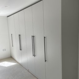 Woodworking - Długa, biała szafa wnękowa z minimalistycznymi, czarnymi uchwytami, widoczna na tle surowego wnętrza w trakcie remontu.