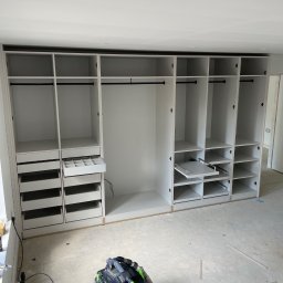 Woodworking - Wnętrze garderoby w trakcie montażu, widoczne wysuwane półki, szuflady na akcesoria i drążki na ubrania, na podłodze leży odkurzacz przemysłowy.