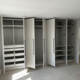 Woodworking - Biała zabudowa meblowa z otwartymi drzwiami, widoczne półki, szuflady i drążki na ubrania, czarne, długie uchwyty, surowy stan podłogi.