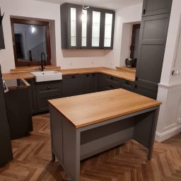 Woodworking - Nowoczesna kuchnia w odcieniach szarości z drewnianym blatem i wyspą, parkiet ułożony w jodełkę, widoczne okno z ciemną ramą i biały zlew.