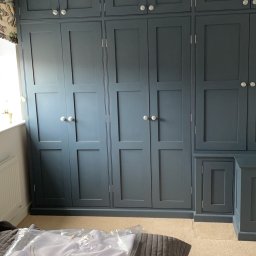 Woodworking - Duża, zabudowana szafa w odcieniach szarości z panelowymi drzwiami i okrągłymi, srebrnymi gałkami, częściowo zasłonięta przez ubranie w plastikowym opakowaniu leżące na łóżku.