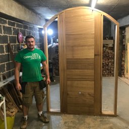 Woodworking - Stolarz prezentuje drewniane drzwi z łukowym wykończeniem, widoczne narzędzia i elementy montażowe w tle.