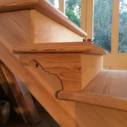 Detal drewnianych schodów z widocznym ozdobnym wykończeniem stopnia i fragmentem okna w tle.