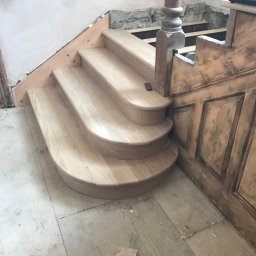 Woodworking - Drewniane schody z zaokrąglonymi stopniami w trakcie montażu, widoczne niedokończone ściany i podłoga, oraz fragment starej, ozdobnej boazerii.