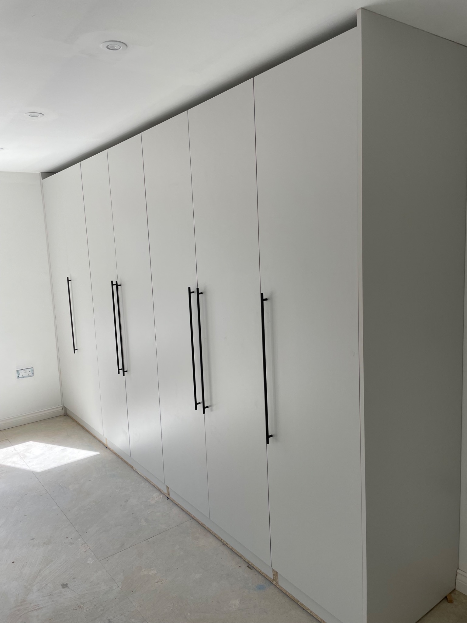 Długa, biała szafa wnękowa z minimalistycznymi, czarnymi uchwytami, widoczna na tle surowego wnętrza w trakcie remontu.