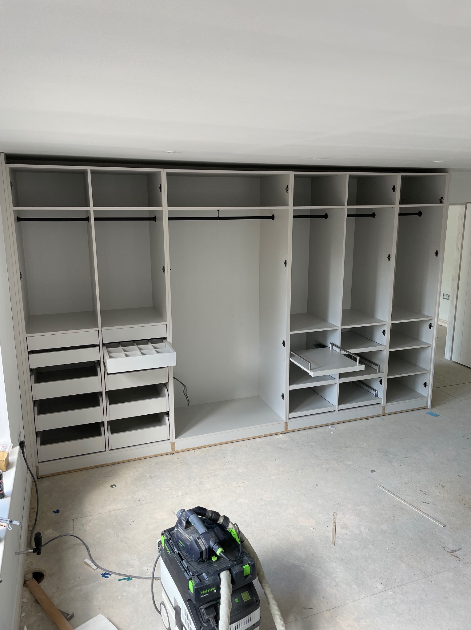 Wnętrze garderoby w trakcie montażu, widoczne wysuwane półki, szuflady na akcesoria i drążki na ubrania, na podłodze leży odkurzacz przemysłowy.