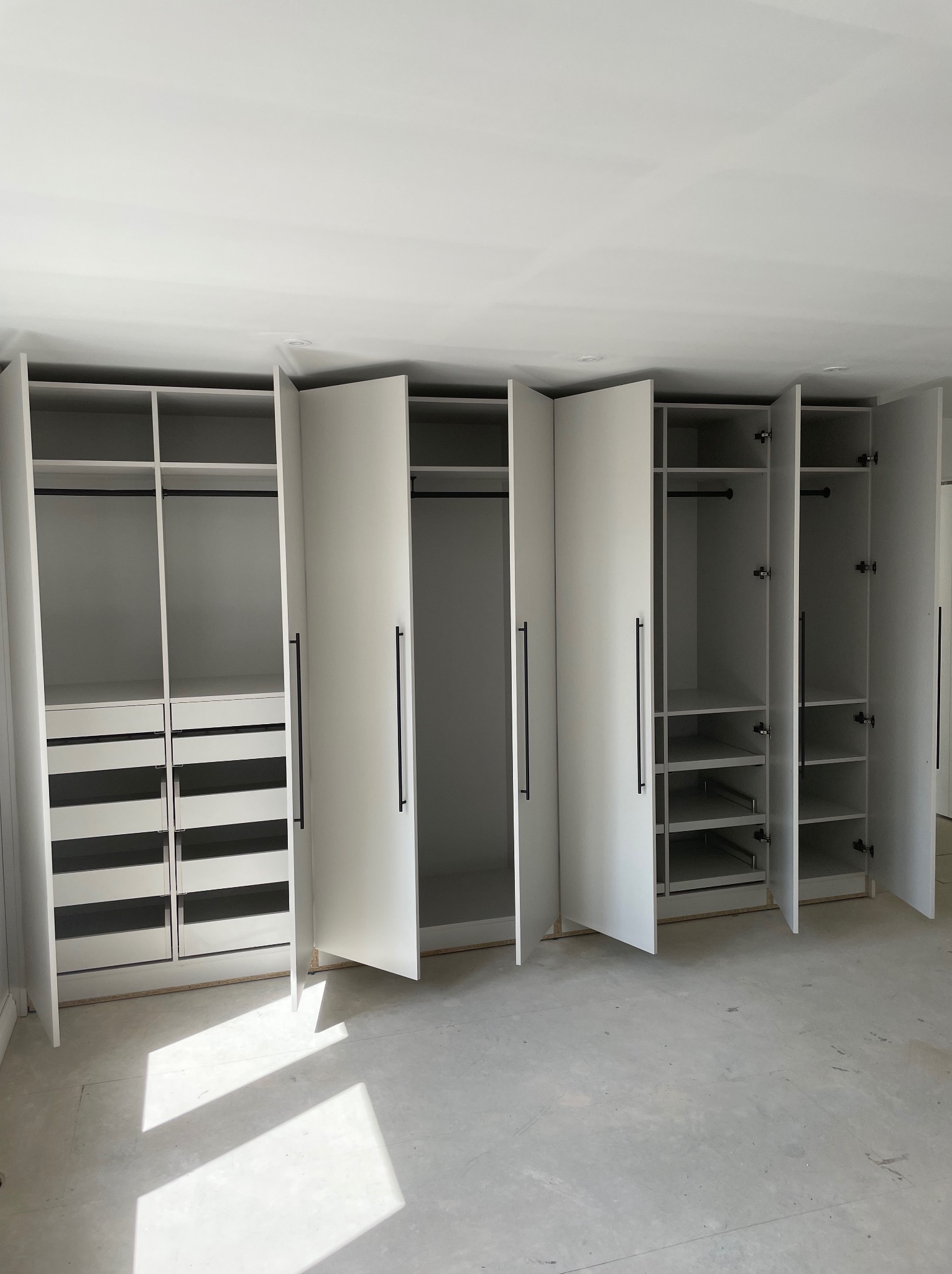 Biała zabudowa meblowa z otwartymi drzwiami, widoczne półki, szuflady i drążki na ubrania, czarne, długie uchwyty, surowy stan podłogi.