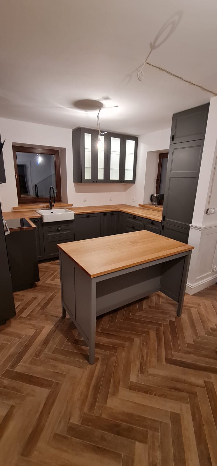 Nowoczesna kuchnia w odcieniach szarości z drewnianym blatem i wyspą, parkiet ułożony w jodełkę, widoczne okno z ciemną ramą i biały zlew.