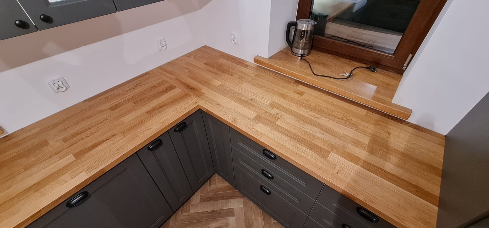Drewniany blat kuchenny w kształcie litery L, szare szafki z czarnymi uchwytami, czajnik elektryczny na parapecie okiennym, parkiet ułożony w jodełkę.