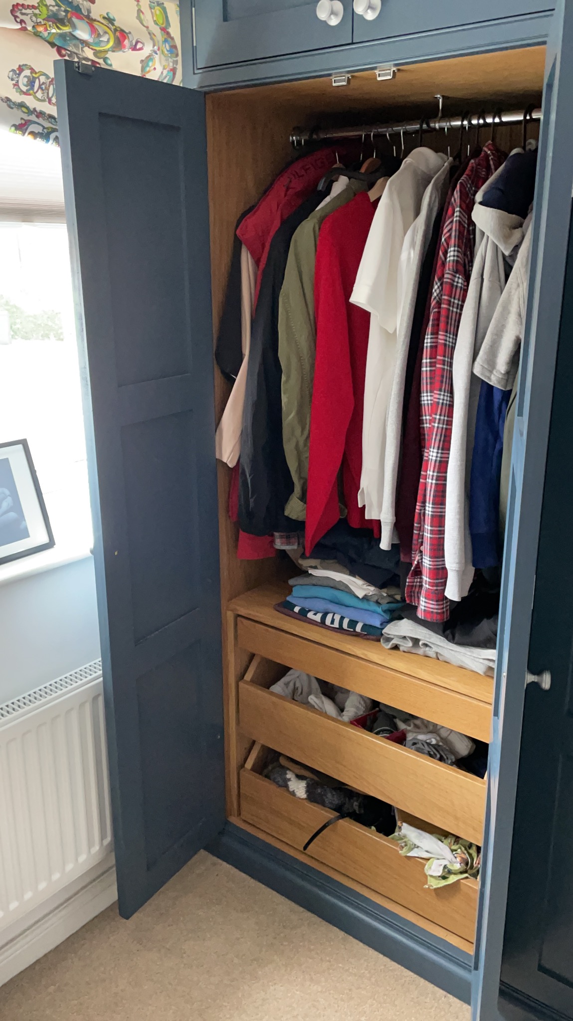 Niebieska szafa z otwartymi drzwiami, widoczne ubrania na wieszakach i w szufladach, wnętrze wykonane z jasnego drewna.