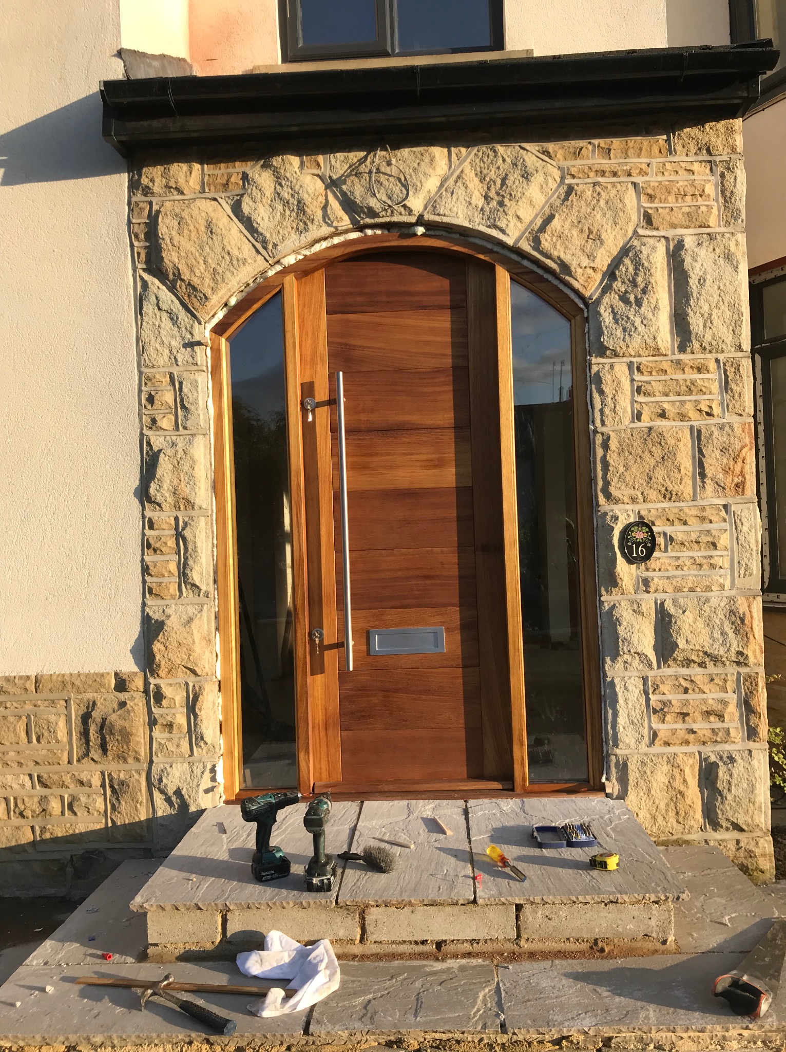 Nowoczesne drzwi wejściowe z drewna egzotycznego, osadzone w kamiennej obudowie łukowej, widoczne narzędzia i akcesoria montażowe na stopniach wejściowych.