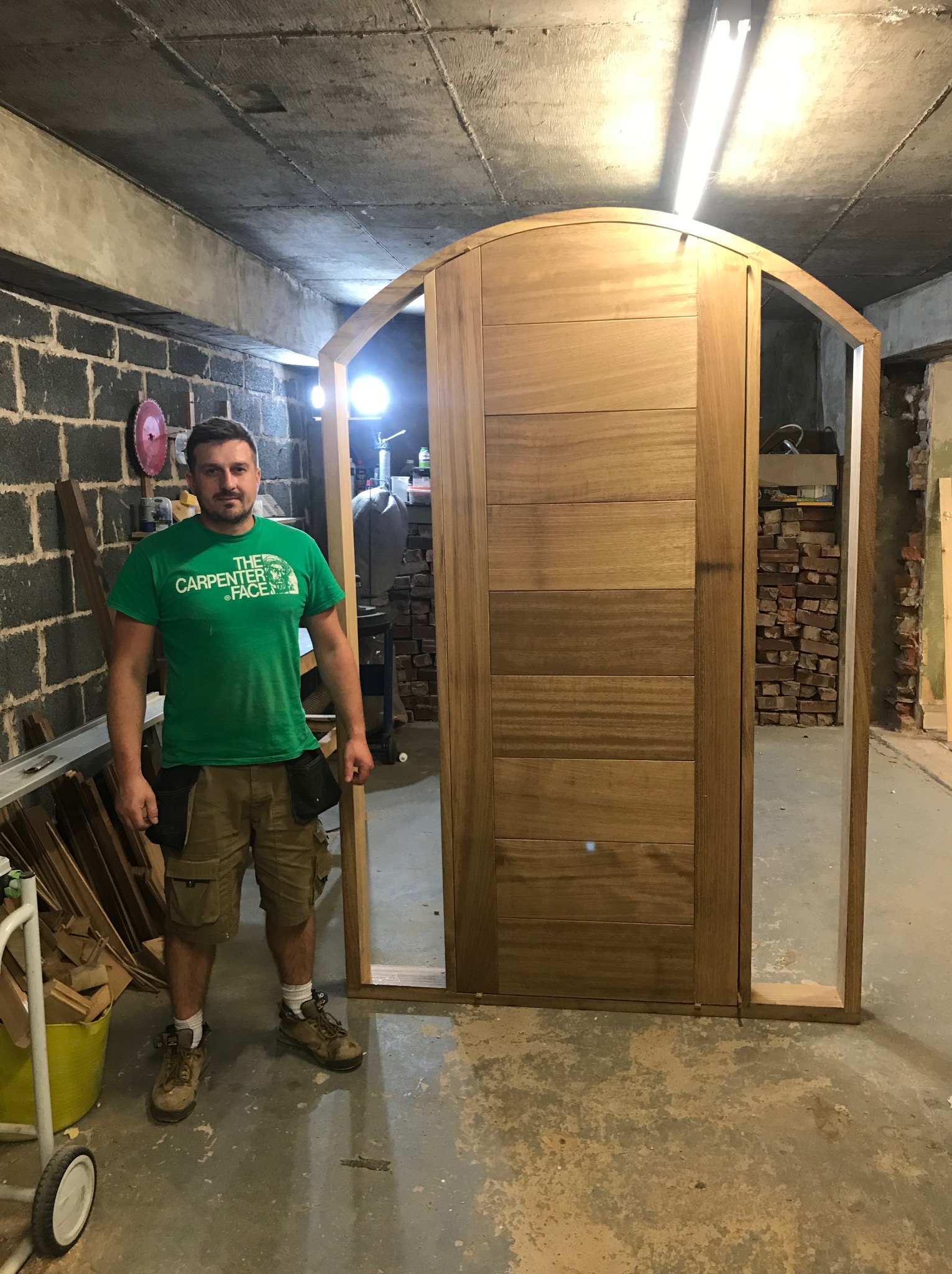 Stolarz prezentuje drewniane drzwi z łukowym wykończeniem, widoczne narzędzia i elementy montażowe w tle.