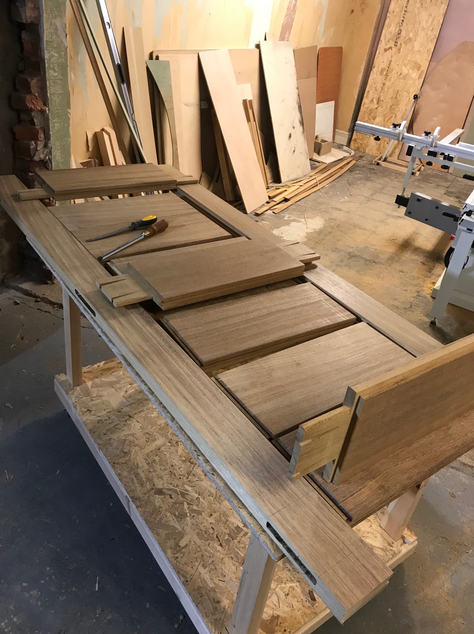 Elementy drewniane do montażu drzwi, leżące na stole warsztatowym w trakcie obróbki, obok leżą dłuto i pilnik.