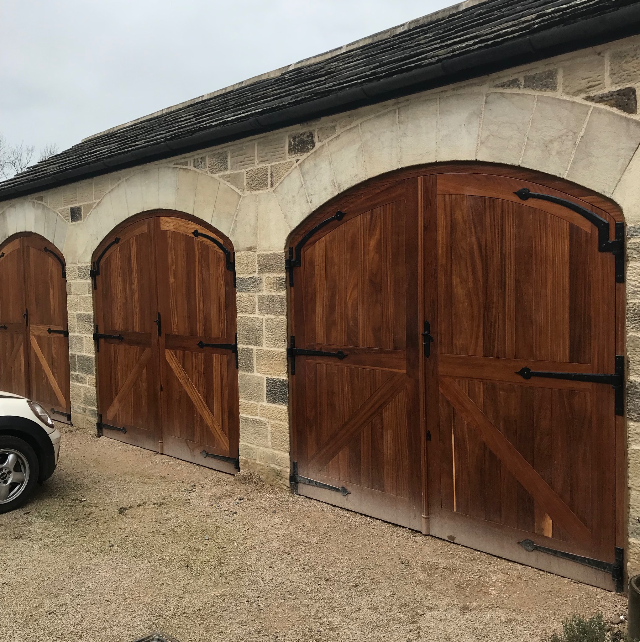 Trzy drewniane, łukowe bramy garażowe z czarnymi okuciami w kamiennym budynku z szarym dachem, częściowo widoczny samochód z lewej strony.