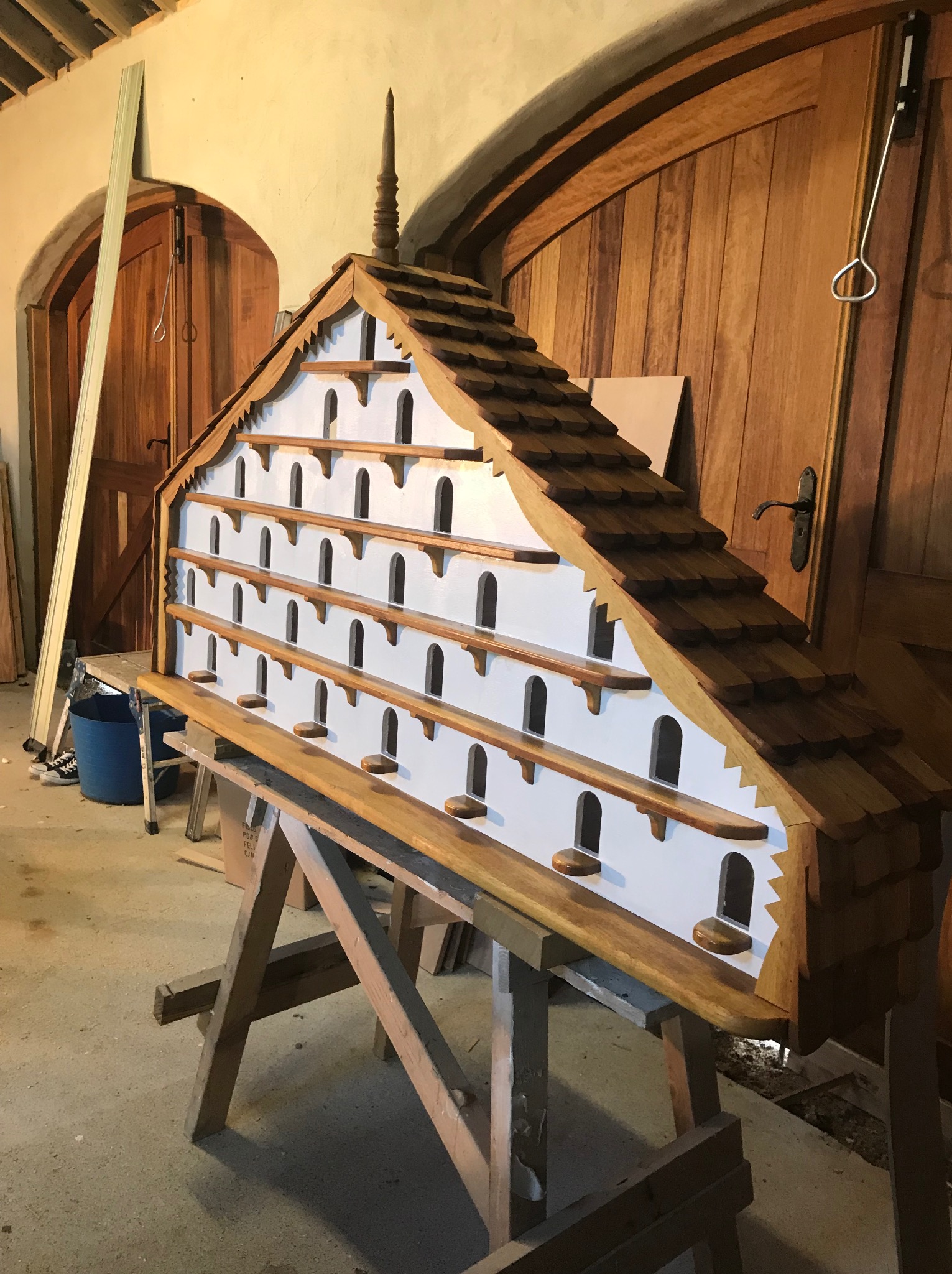 Duża, drewniana budka dla ptaków w kształcie domu z wieloma otworami, umieszczona na drewnianym stojaku w warsztacie, z widocznymi drzwiami z drewna w tle.