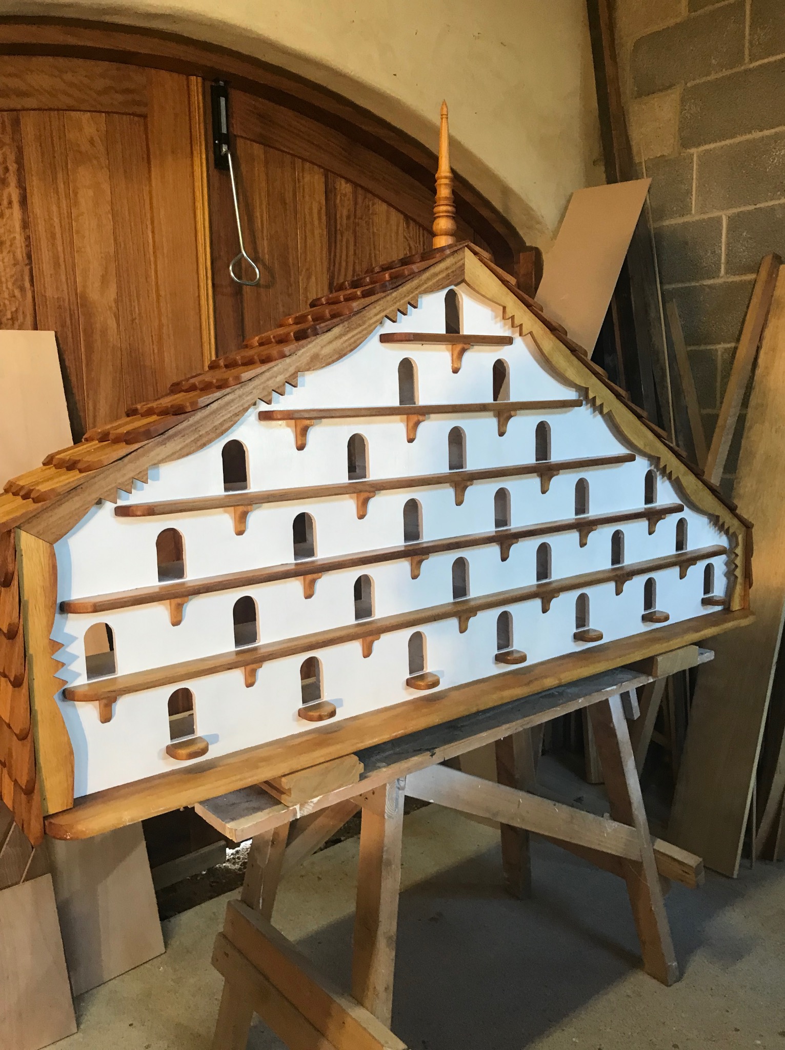 Drewniana konstrukcja domku dla ptaków, surowy stan wykończenia, widoczne elementy stolarskie, umieszczona na drewnianych kozłach w warsztacie.