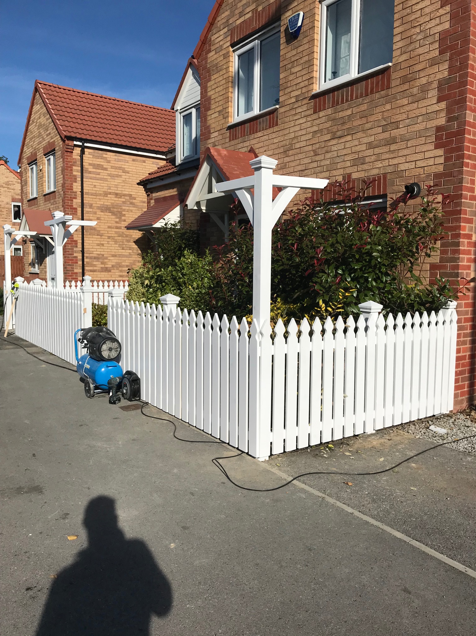Białe, drewniane ogrodzenie sztachetowe przed domem z czerwonej cegły, obok stoi niebieski kompresor malarski, cień osoby na chodniku.