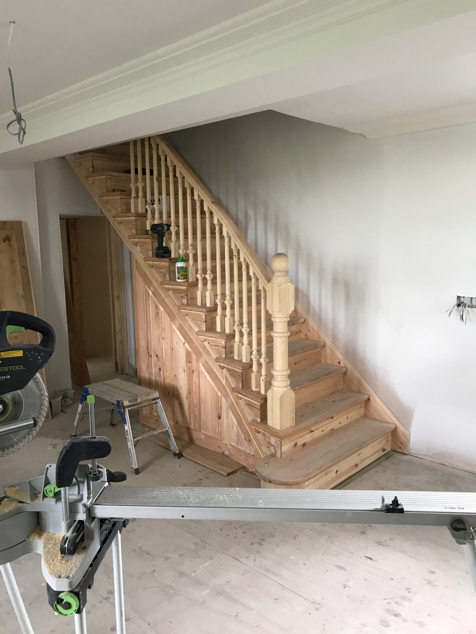 Montaż drewnianych schodów z balustradą w surowym stanie, widoczna piła stołowa i narzędzia, wnętrze w trakcie remontu.
