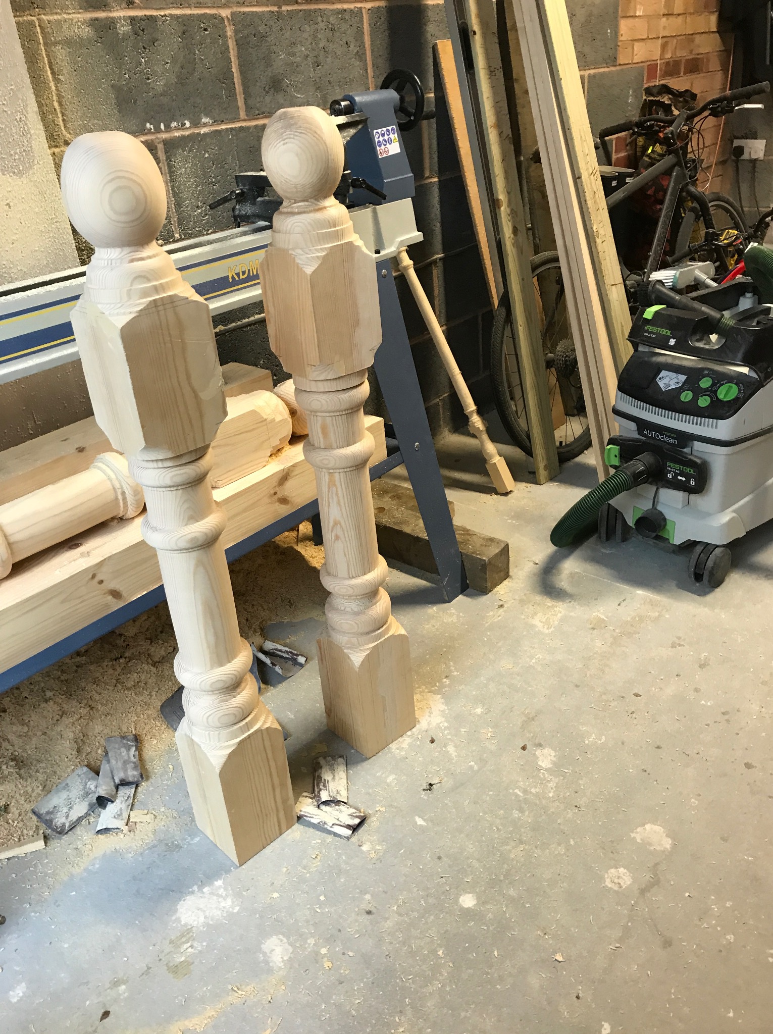 Dwie surowe, drewniane kolumny balustrady z kulistymi zakończeniami, obok tokarki do drewna i odkurzacza przemysłowego, w warsztacie.