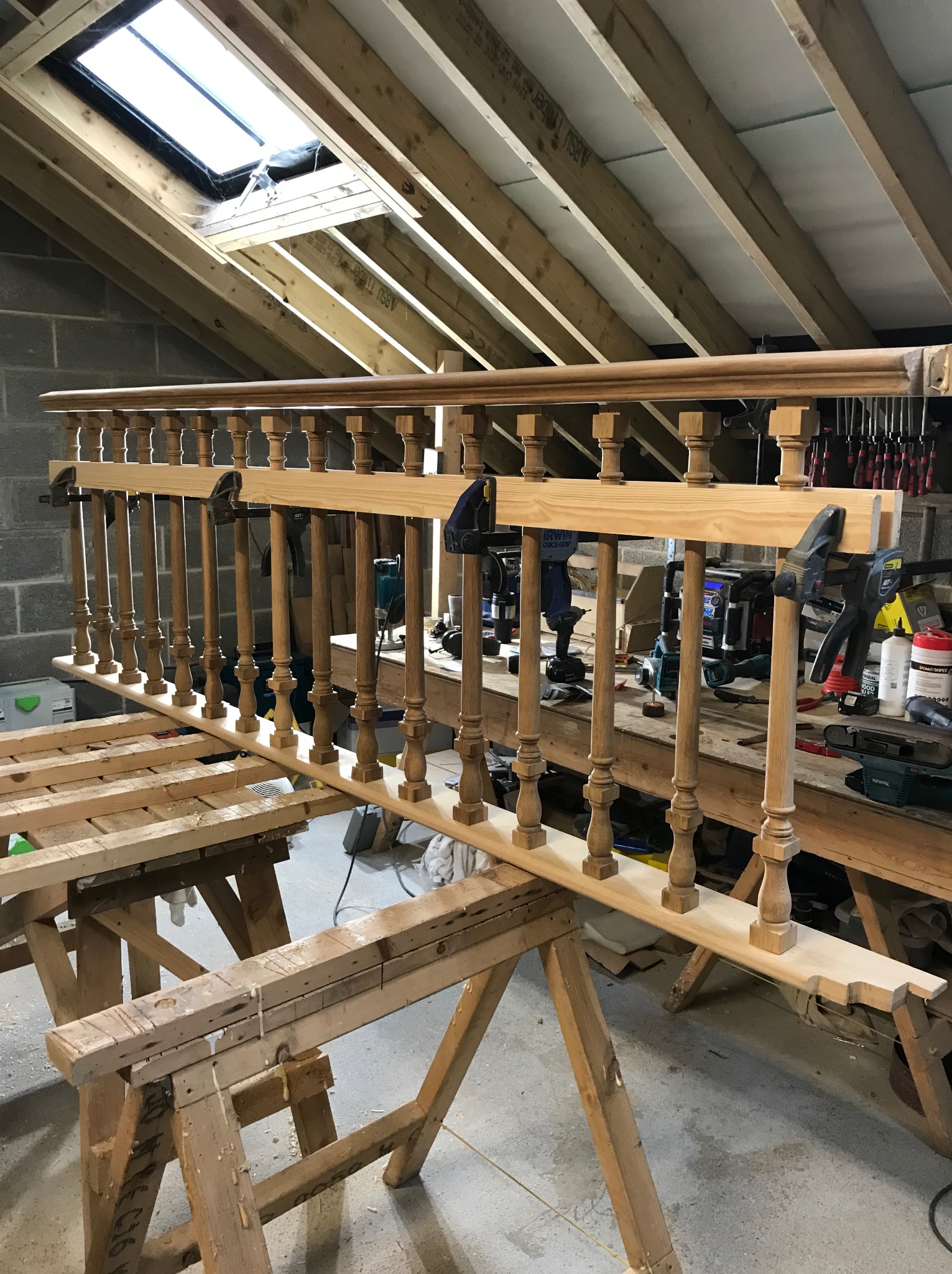 Drewniana balustrada w trakcie montażu w warsztacie, zabezpieczona klamrami stolarskimi, widoczne narzędzia i akcesoria, surowe drewno i fragment dachu z oknem.