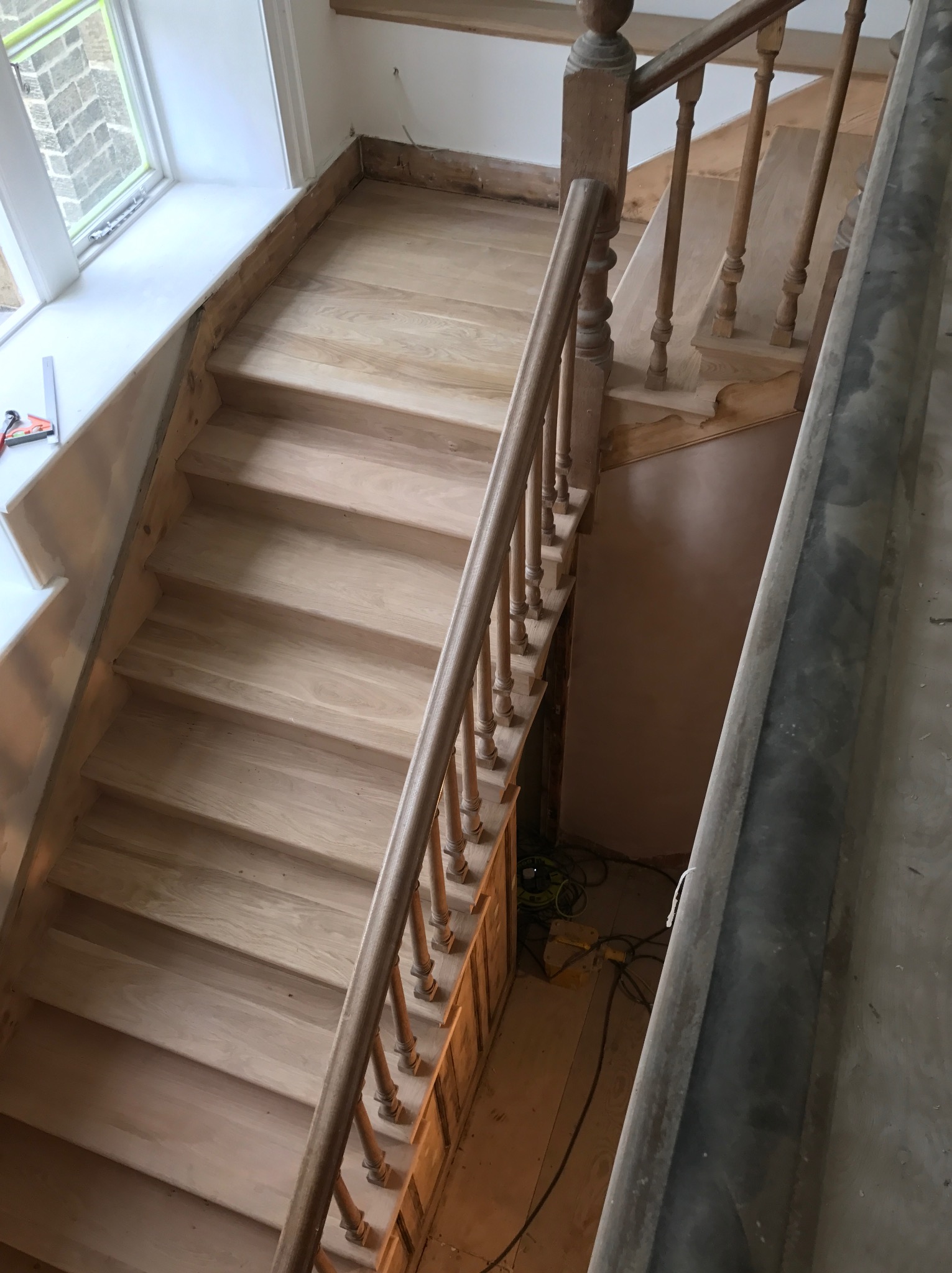 Jasne drewniane schody z balustradą w trakcie montażu, widoczne niedokończone wykończenia i narzędzia w tle.