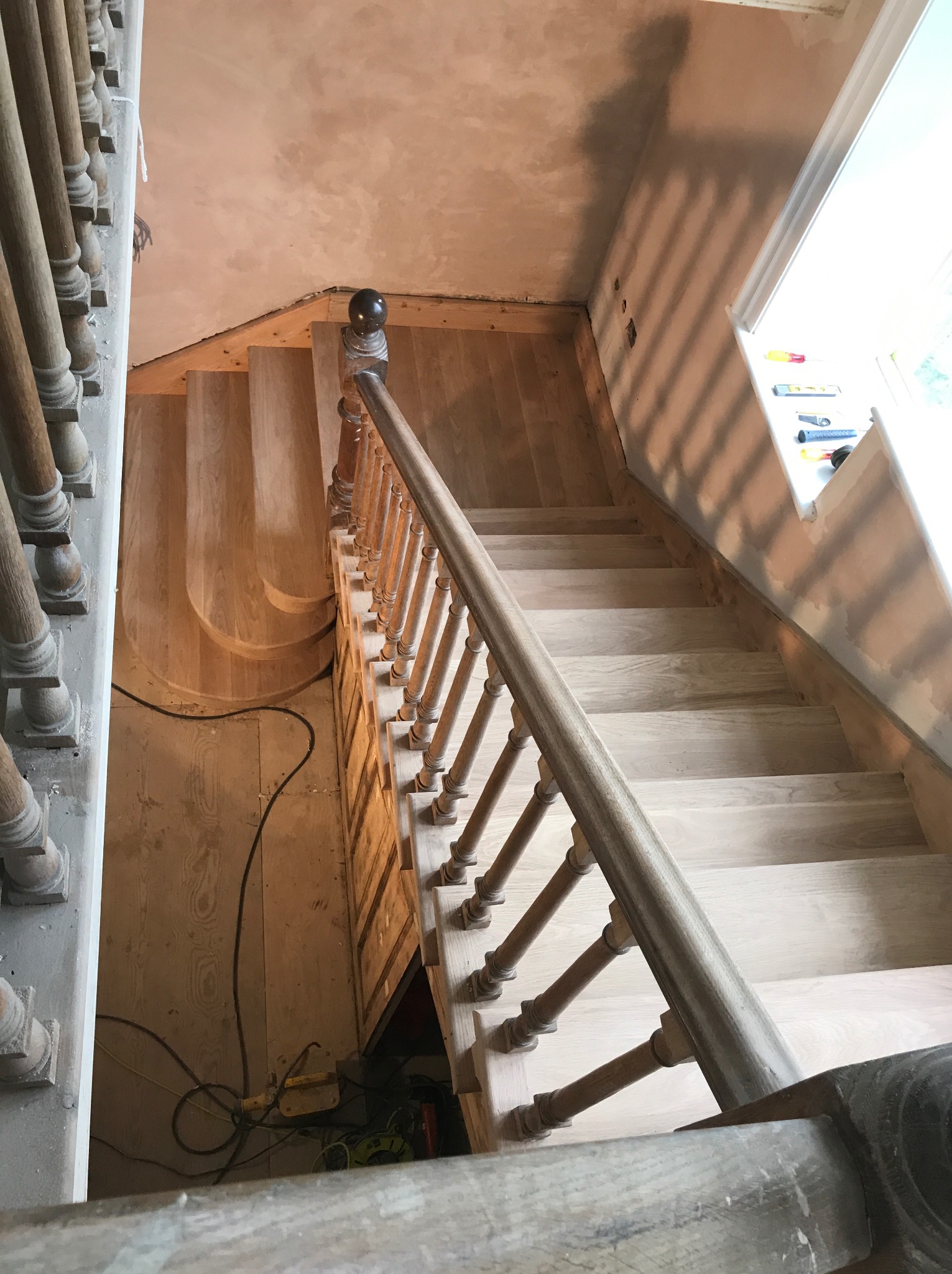 Wnętrze domu w trakcie remontu schodów drewnianych z balustradą, widoczne narzędzia i przewody elektryczne, surowy stan wykończenia ścian.