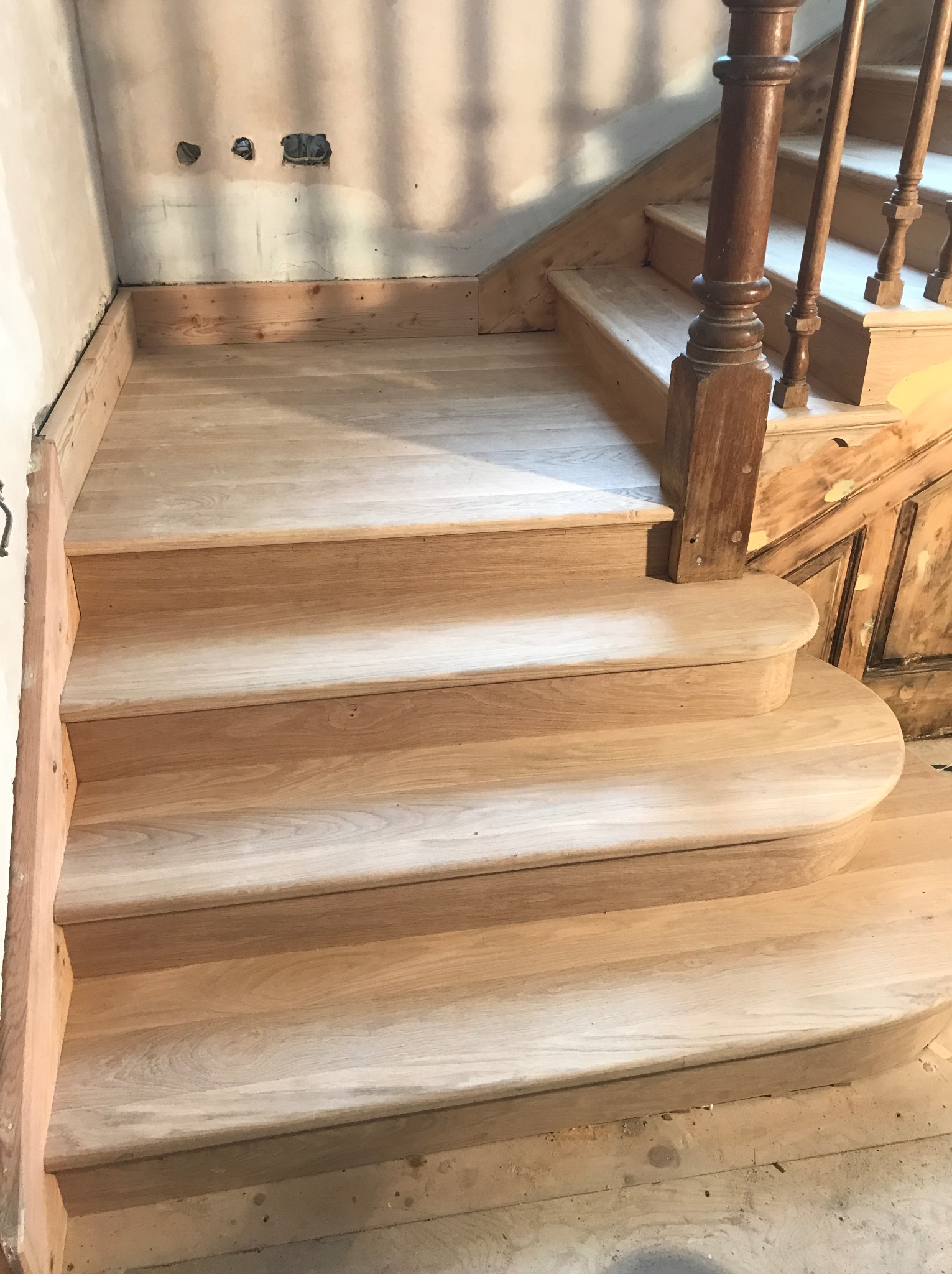 Drewniane schody w trakcie montażu, widoczne surowe ściany i przygotowane podłoże, balustrada z ciemnego drewna.