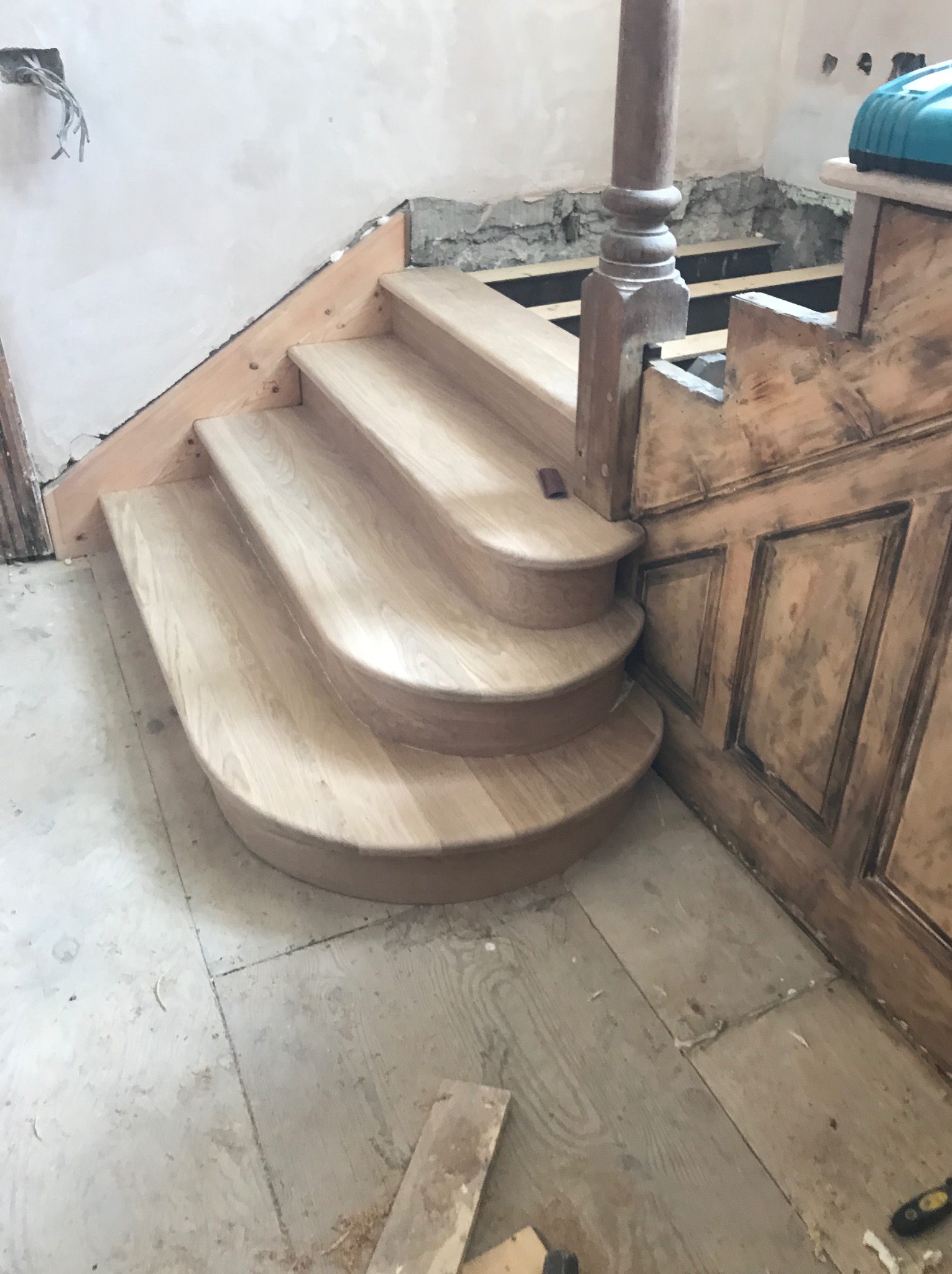 Drewniane schody z zaokrąglonymi stopniami w trakcie montażu, widoczne niedokończone ściany i podłoga, oraz fragment starej, ozdobnej boazerii.