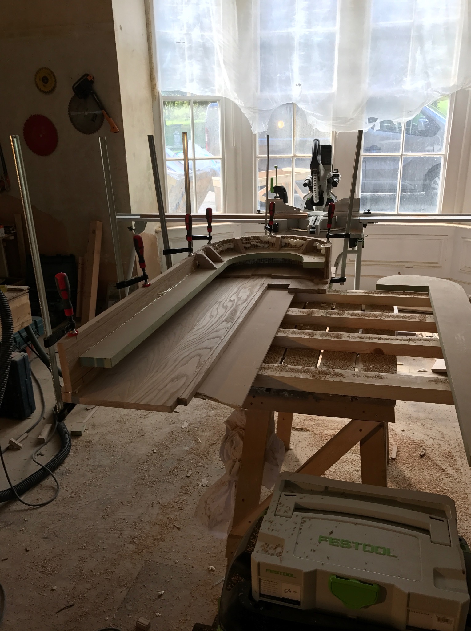 Warsztat stolarski: Fragment drewnianej konstrukcji z przyklejonym elementem, zabezpieczony ściskami, piła ukośnica w tle, narzędzia na ścianie, podłoga pokryta wiórami.