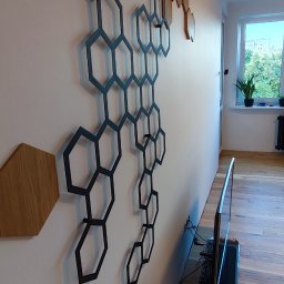 Remont Szczecin - Nowoczesna dekoracja ścienna z heksagonów w salonie. Czarne, metalowe elementy tworzą geometryczny wzór na białej ścianie, obok drewnianych akcentów i roślin.