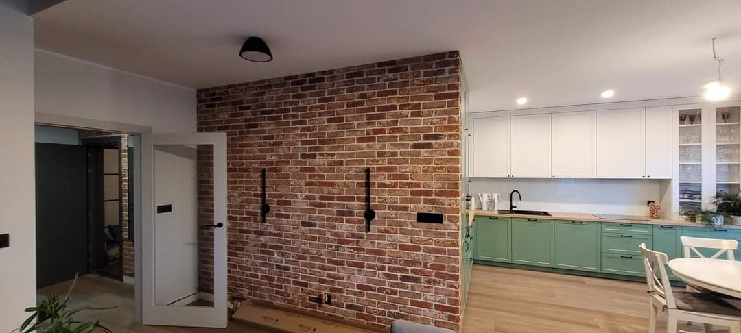 Ceglana ściana z dekoracyjnymi elementami w nowoczesnym wnętrzu. Widoczna kuchnia z zielonymi szafkami i białymi górnymi szafkami. Uwagę przykuwa kontrast materiałów.