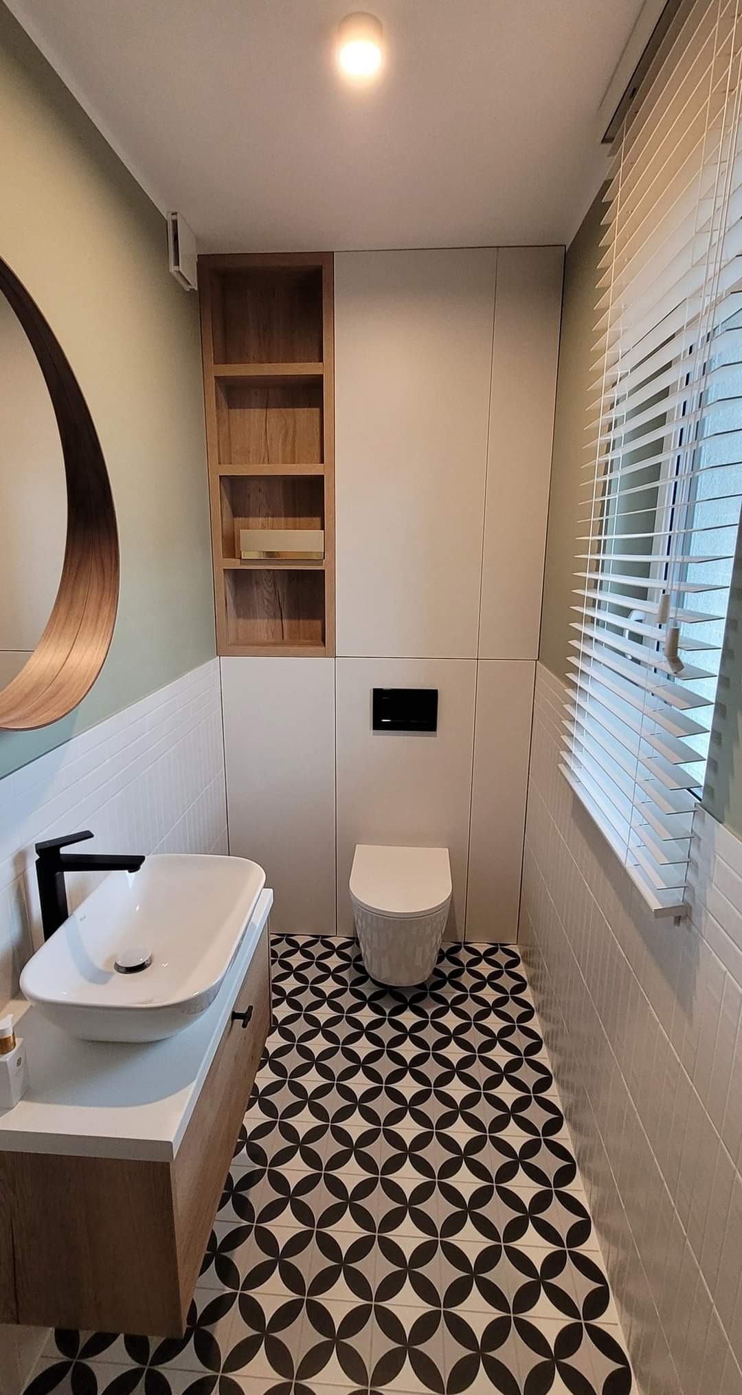 Nowoczesna łazienka z umywalką nablatową, geometryczną podłogą i zabudową meblową w odcieniach bieli i drewna. Okrągłe lustro i żaluzje okienne.