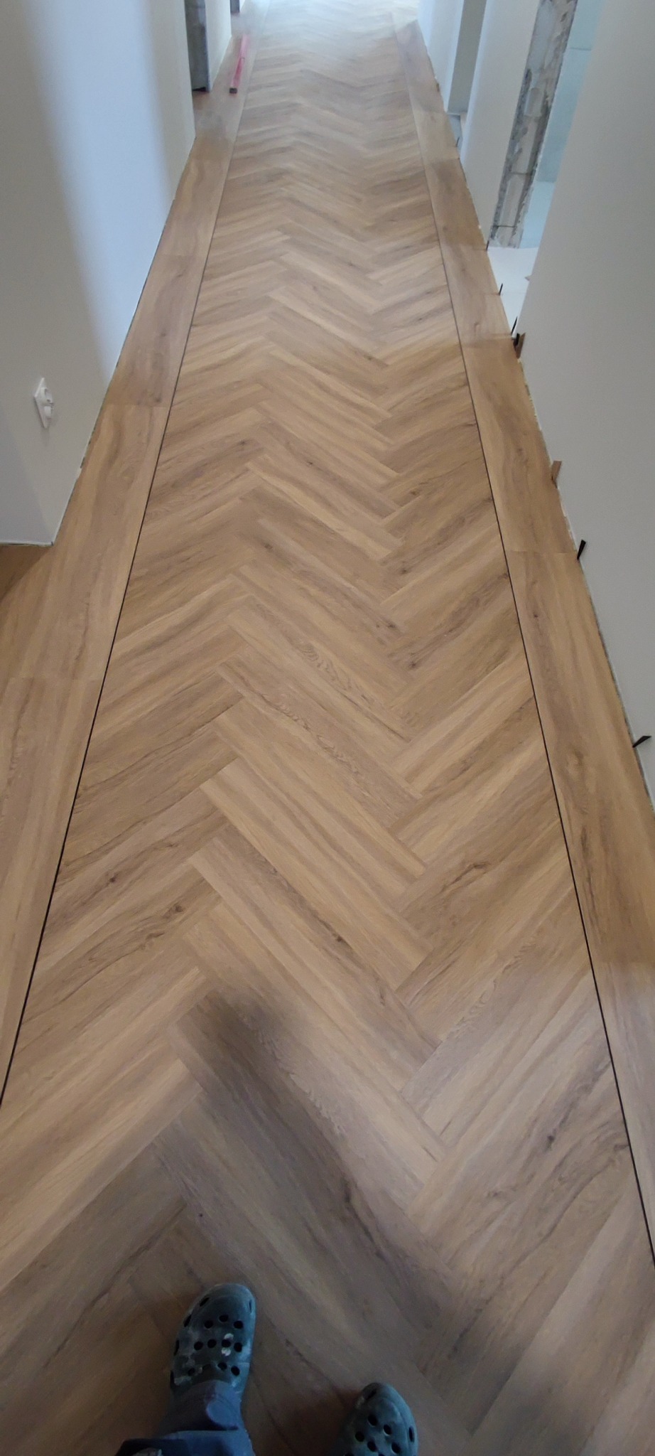Ukończona podłoga z paneli ułożonych we wzór jodełki w korytarzu. Widoczne listwy przypodłogowe i fragment butów osoby wykonującej zdjęcie.