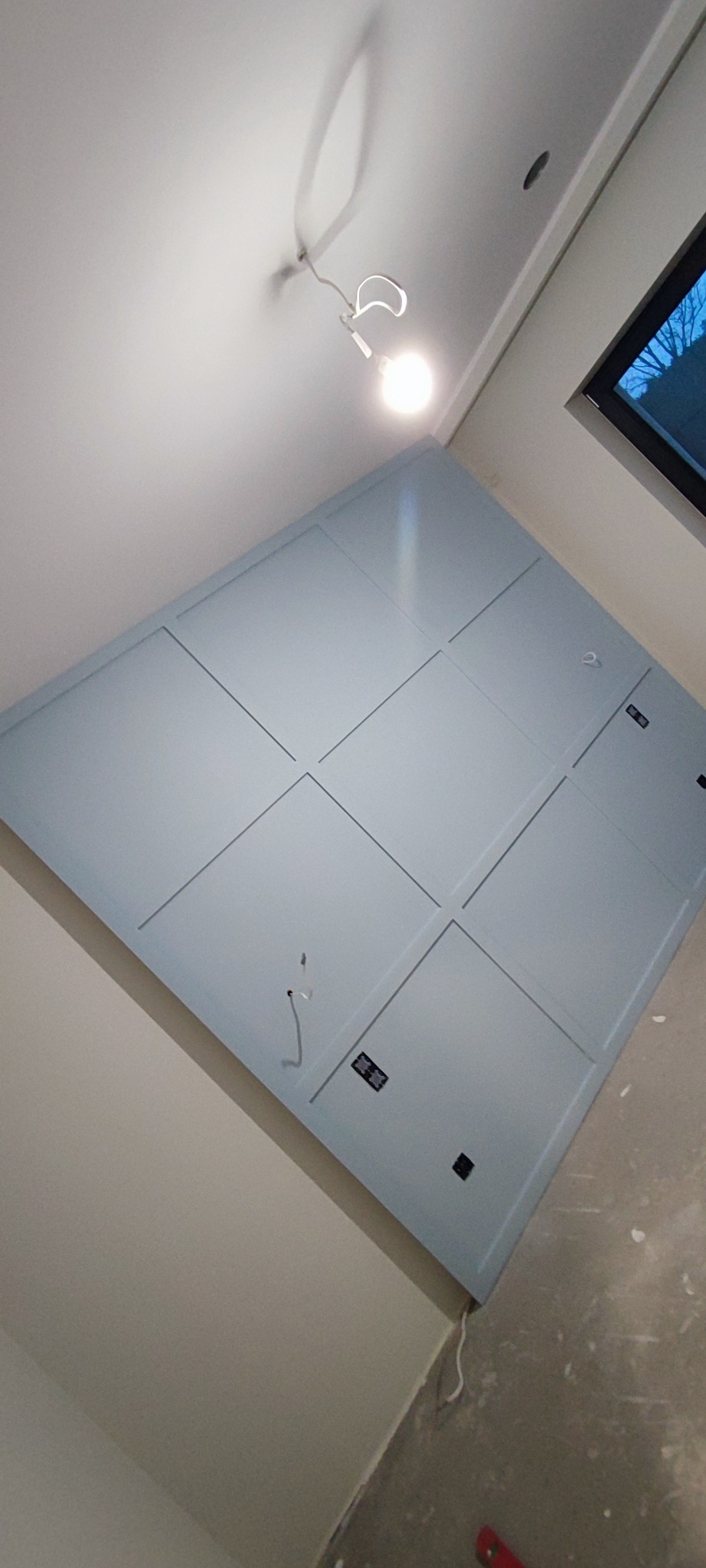 Ściana z płyty karton-gips pomalowana na szaro z dekoracyjnymi listwami i widocznymi puszkami elektrycznymi, oświetlona lampą sufitową.