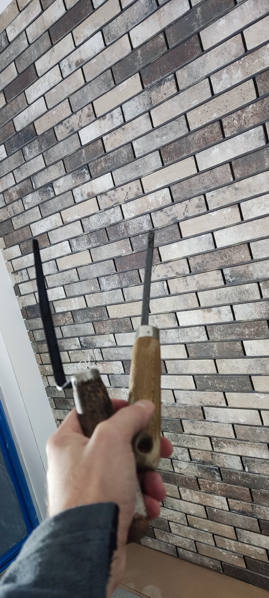 Wykończona ściana z dekoracyjnej cegły w odcieniach beżu i brązu, z narzędziami glazurniczymi w dłoni, sugerującymi etap fugowania. Detal wykończenia wnętrza.