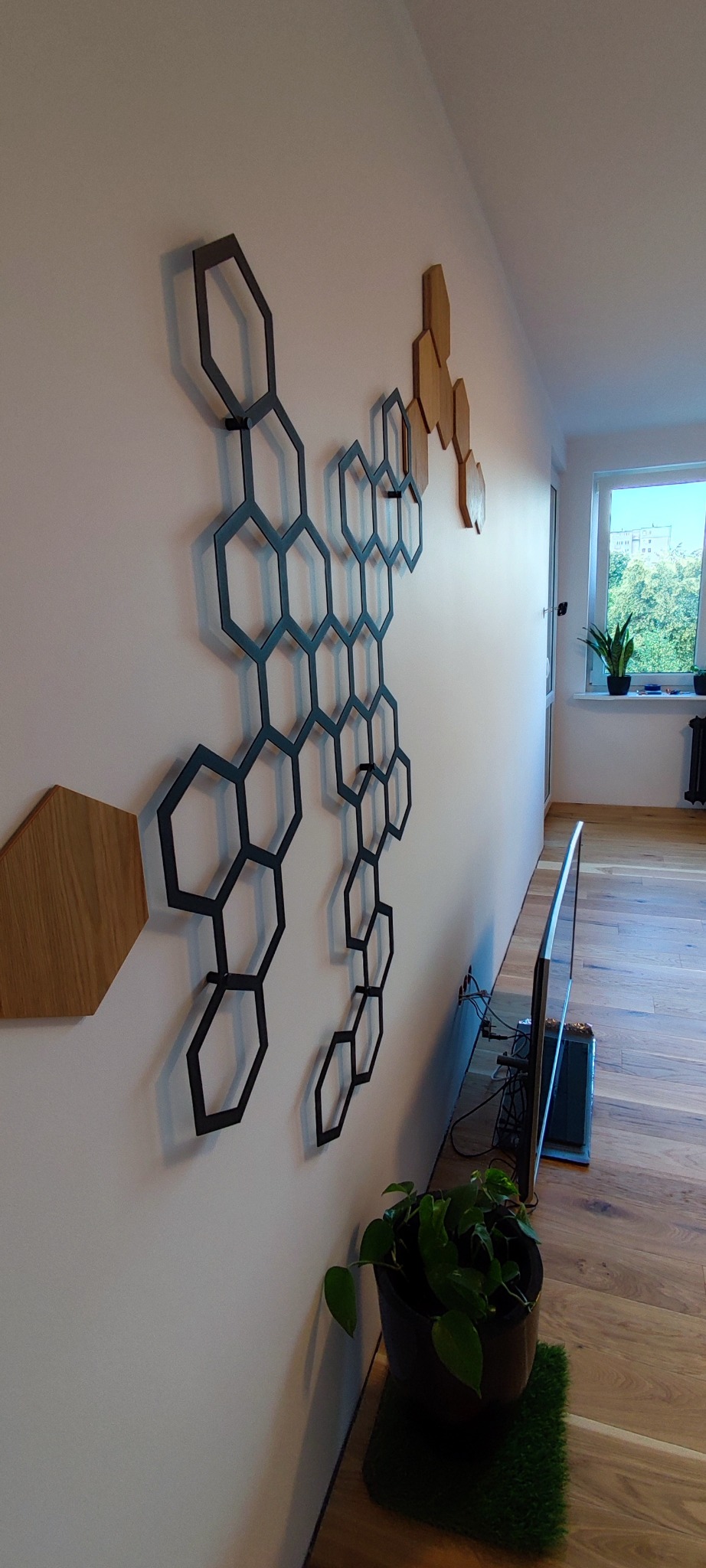 Nowoczesna dekoracja ścienna z heksagonów w salonie. Czarne, metalowe elementy tworzą geometryczny wzór na białej ścianie, obok drewnianych akcentów i roślin.