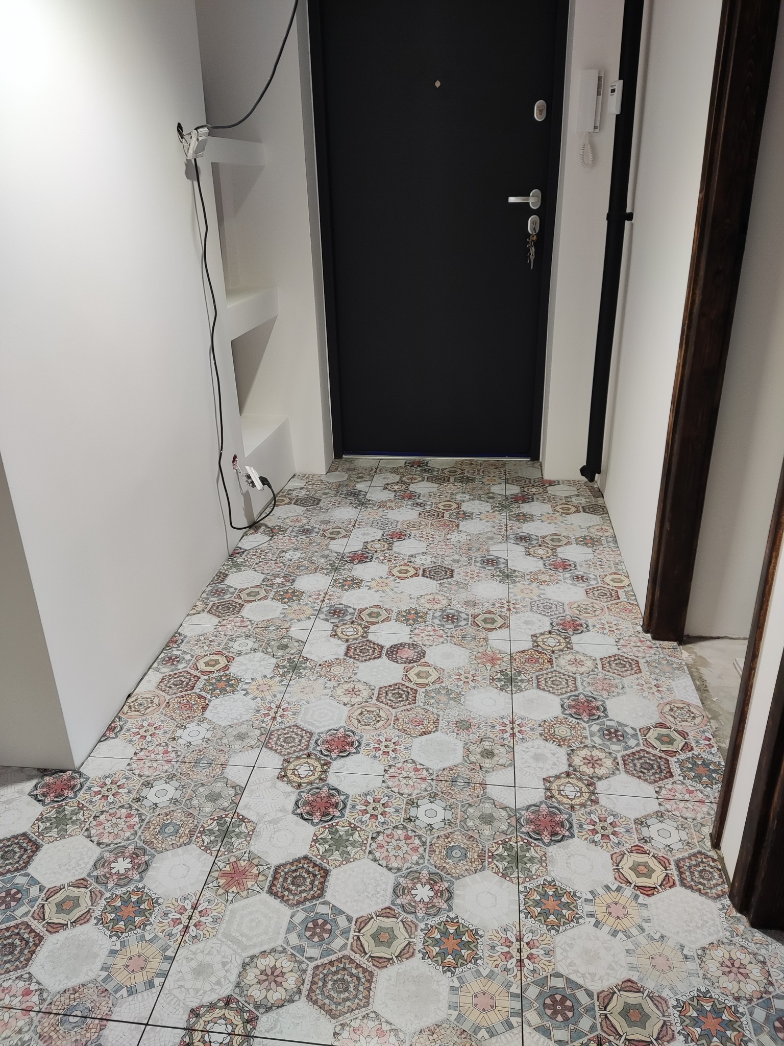 Wnętrze przedpokoju z heksagonalną, wzorzystą posadzką z płytek. Czarna, nowoczesna futryna drzwi wejściowych. Białe ściany i minimalistyczne półki wnękowe.