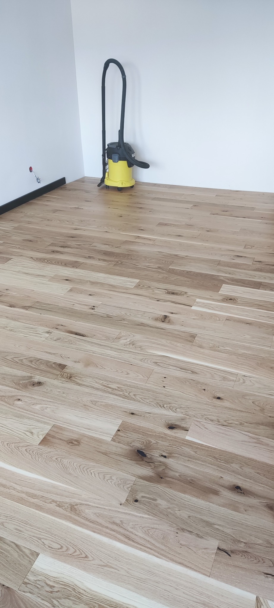 Jasne, drewniane panele podłogowe ułożone w nowym pokoju, widoczne z perspektywy podłogi, z odkurzaczem Karcher w rogu. Listwy przypodłogowe.