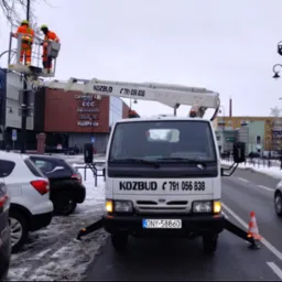 Biały samochód z podnośnikiem koszowym firmy Kozbud, z dwoma pracownikami w pomarańczowych kombinezonach na platformie, ustawiony na ulicy w pobliżu budynków handlowych w pochmurny dzień.