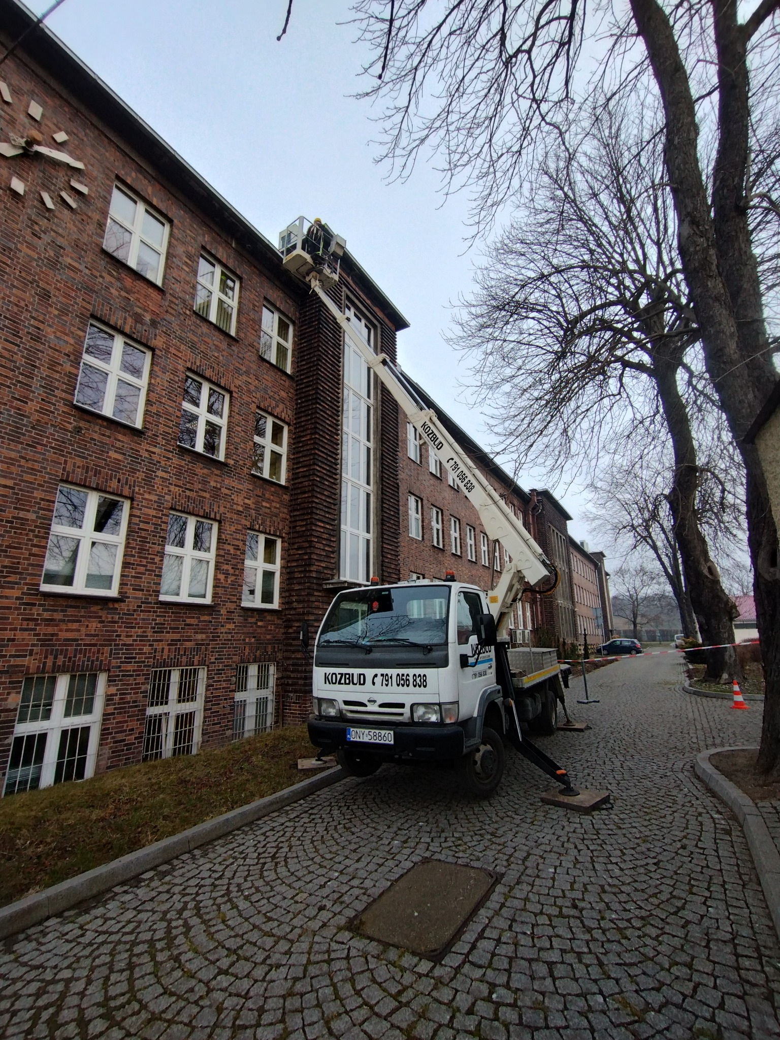 Ciężarówka z wysięgnikiem marki Kozbud ustawiona na brukowanej drodze przed ceglanym budynkiem z zegarem na elewacji, pracownik w koszu na wysokości okien.