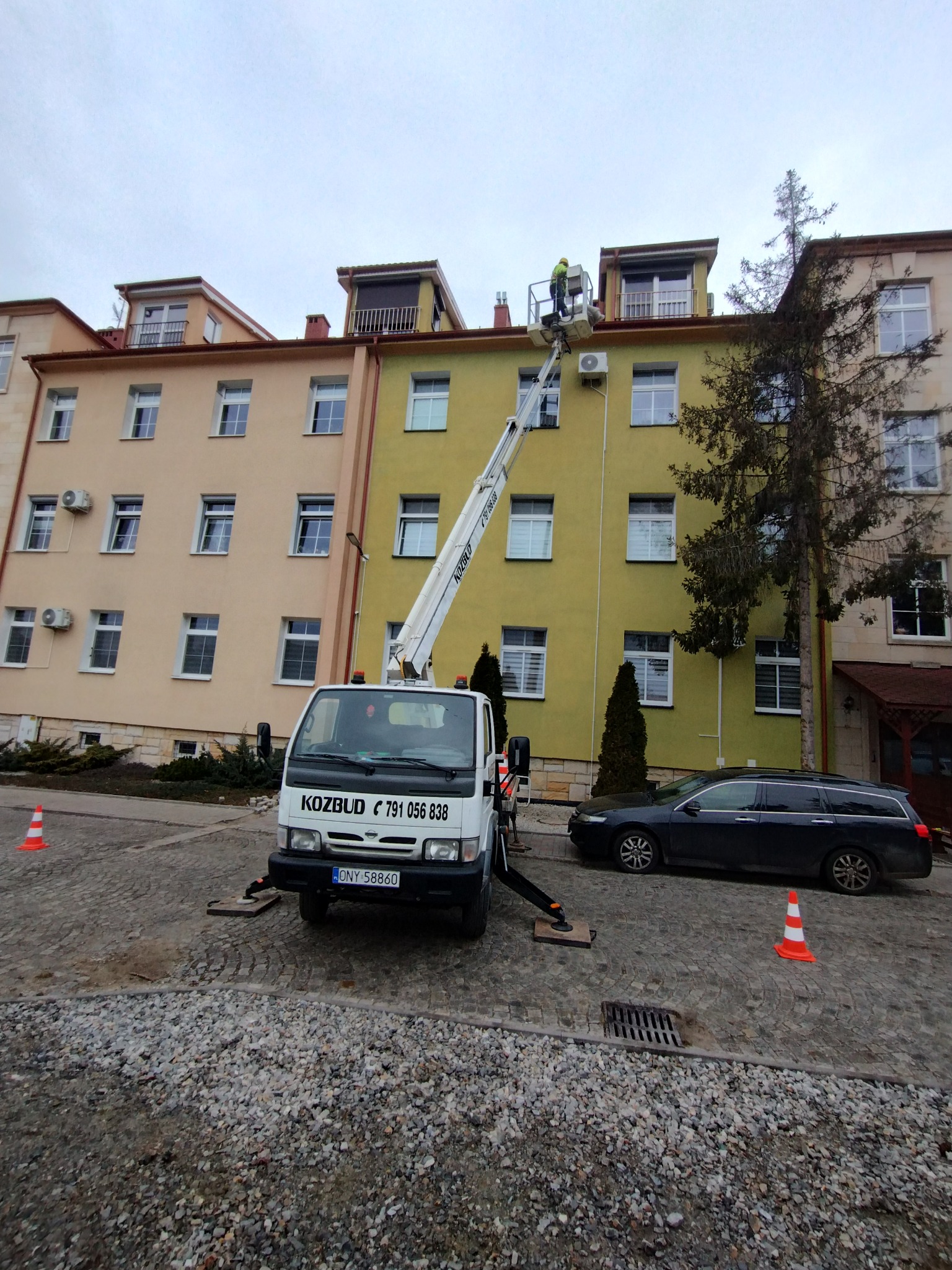 Pracownik w żółtym kasku na podnośniku koszowym przy oknie budynku o żółtej elewacji, na pierwszym planie zaparkowany czarny samochód kombi i biała ciężarówka z wysięgnikiem na brukowanym podłożu.