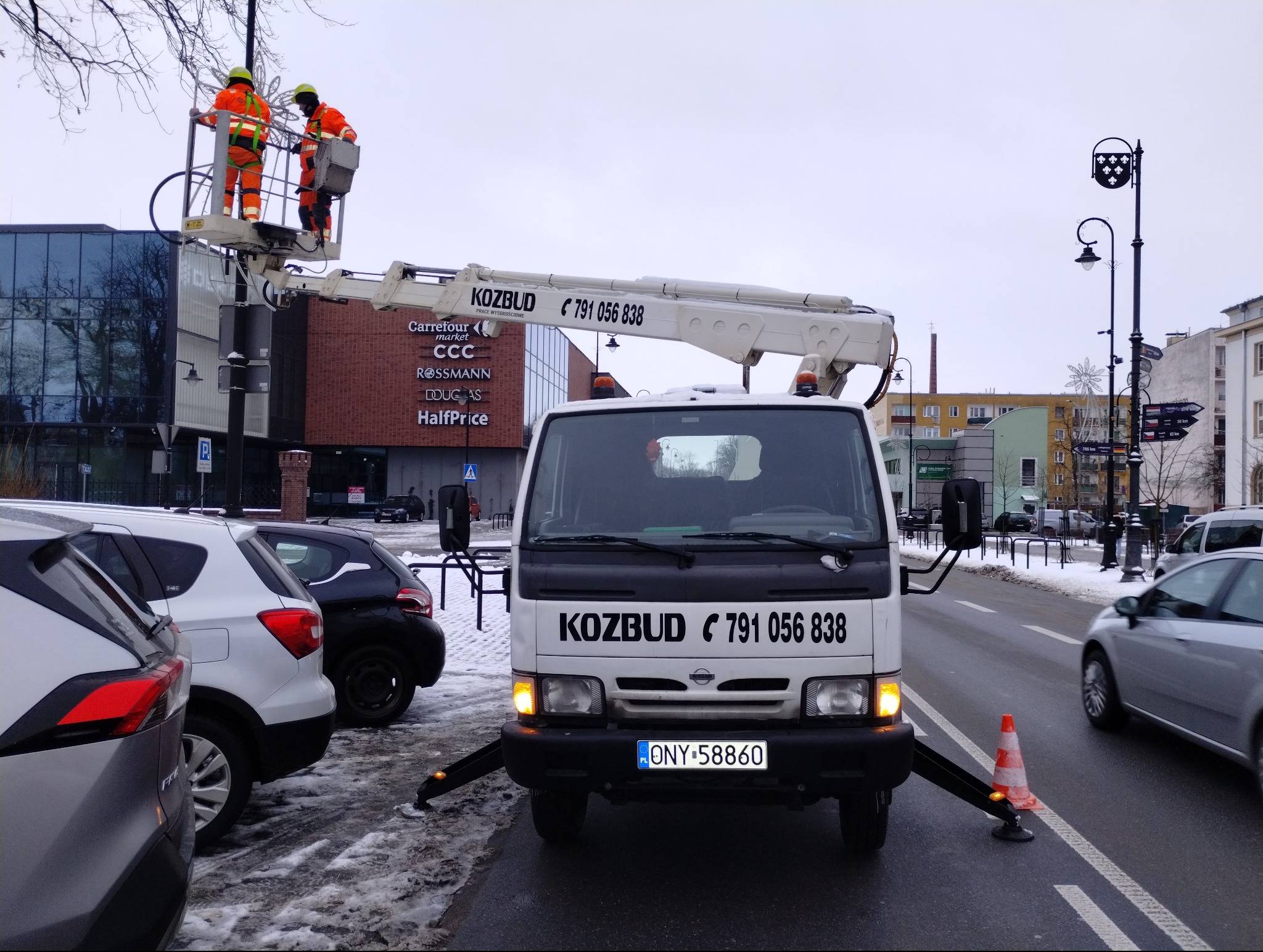 Biały samochód z podnośnikiem koszowym firmy Kozbud, z dwoma pracownikami w pomarańczowych kombinezonach na platformie, ustawiony na ulicy w pobliżu budynków handlowych w pochmurny dzień.