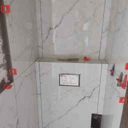 DOB -MAX PAWEŁ DOBOSZ - Białe płytki imitujące marmur z czerwonymi klipsami poziomującymi, zamontowane w łazience. Widoczne otwory na instalacje i odbicie osoby robiącej zdjęcie.