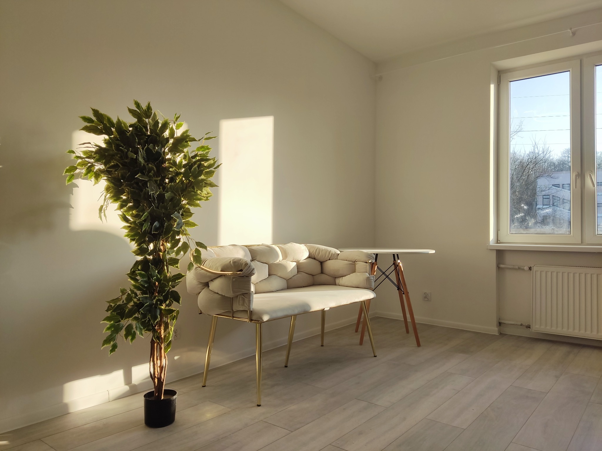 Jasne wnętrze z drewnianą podłogą, na której stoi designerska ławka z beżową tapicerką w kształcie plastra miodu i złotymi nogami, obok zielona roślina w czarnej doniczce i mały stolik z białym...