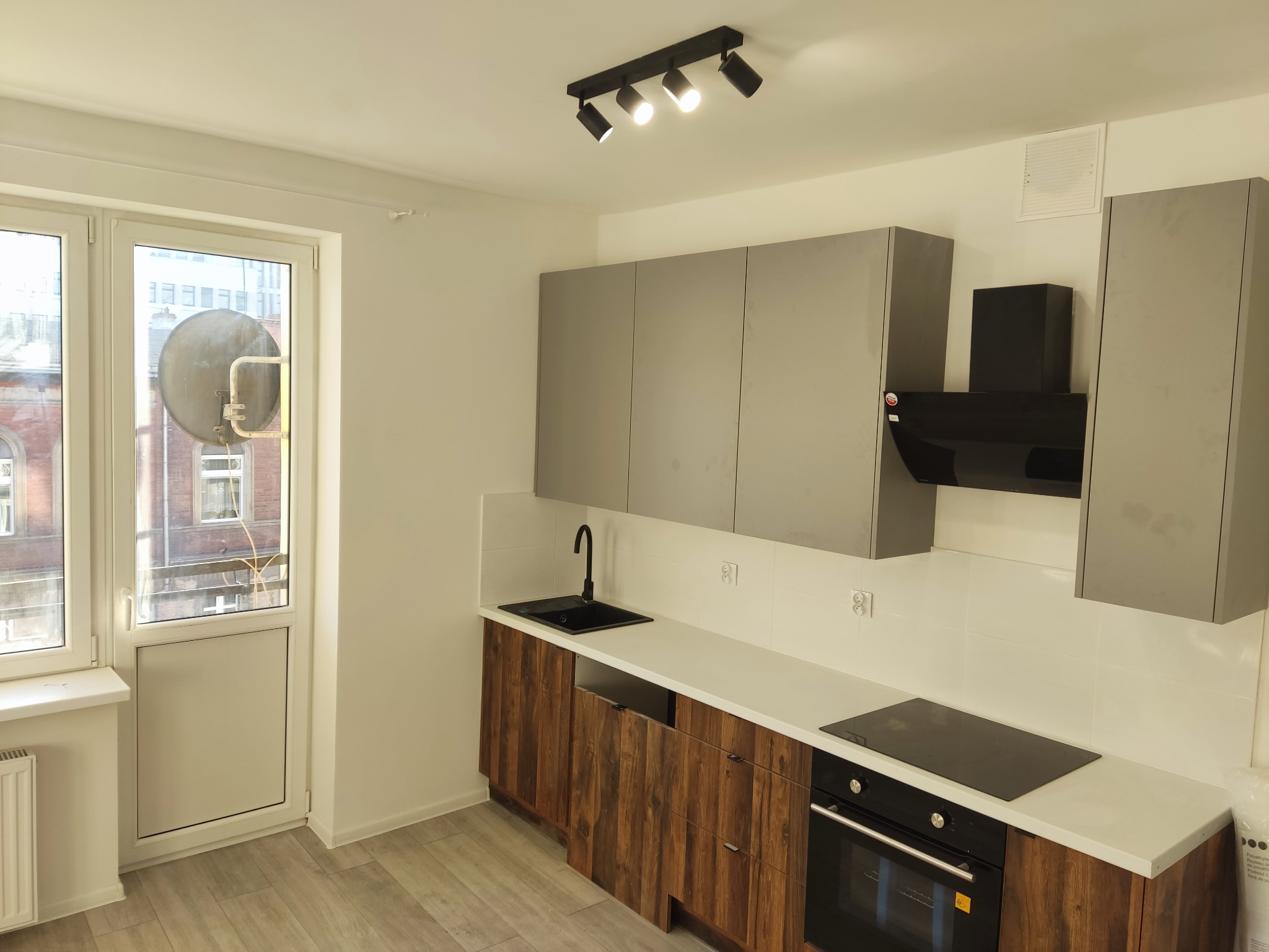 Nowoczesna kuchnia z szarymi szafkami górnymi i drewnopodobnymi dolnymi, czarnym zlewem i płytą indukcyjną, oświetlona reflektorami, z widokiem na budynek przez okno balkonowe.