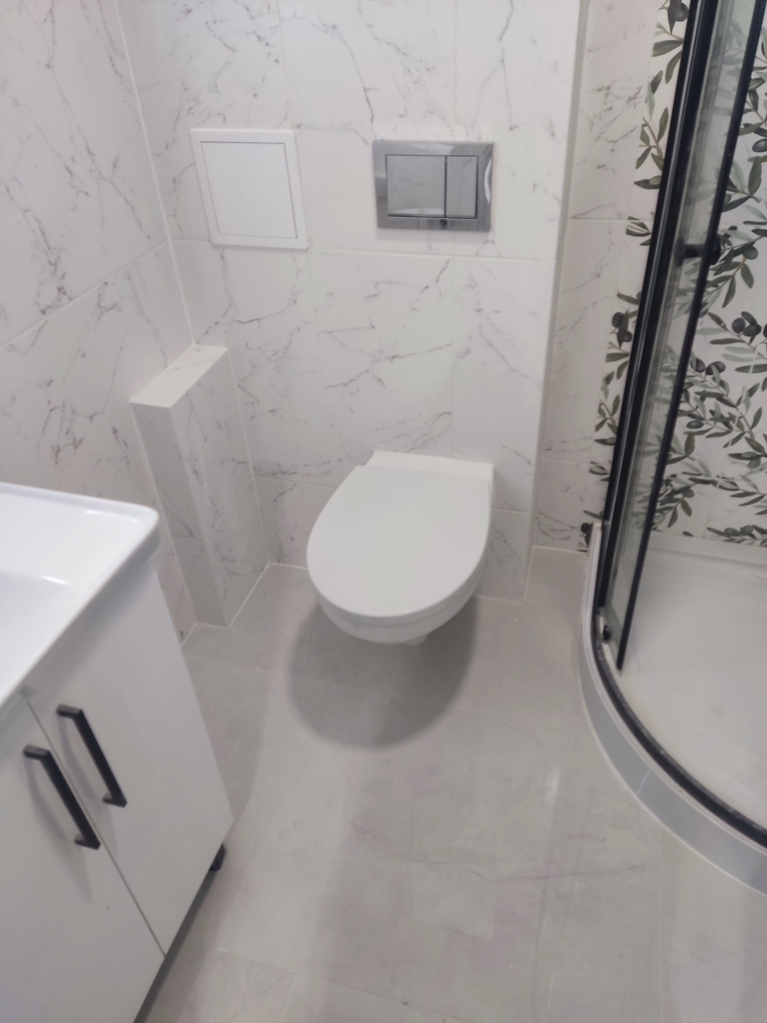 Nowoczesna łazienka z białymi marmurkowymi płytkami, podwieszaną toaletą z chromowanym przyciskiem spłukiwania, białą szafką pod umywalką z czarnymi uchwytami i kabiną prysznicową z wzorem liści...