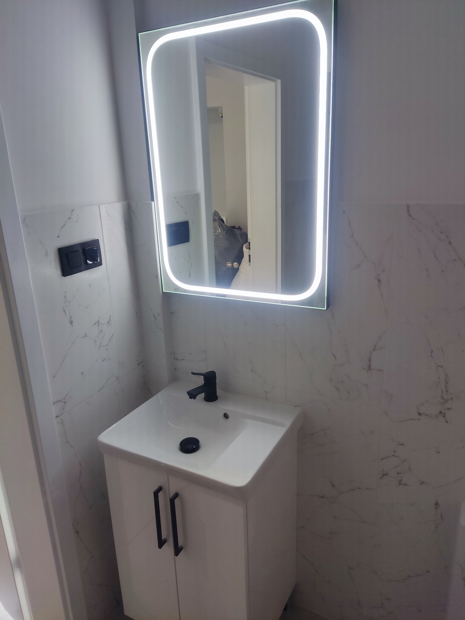 Nowoczesna łazienka z umywalką z szafką w kolorze białym, czarną armaturą i podświetlanym lustrem LED na tle marmurowych płytek ściennych.