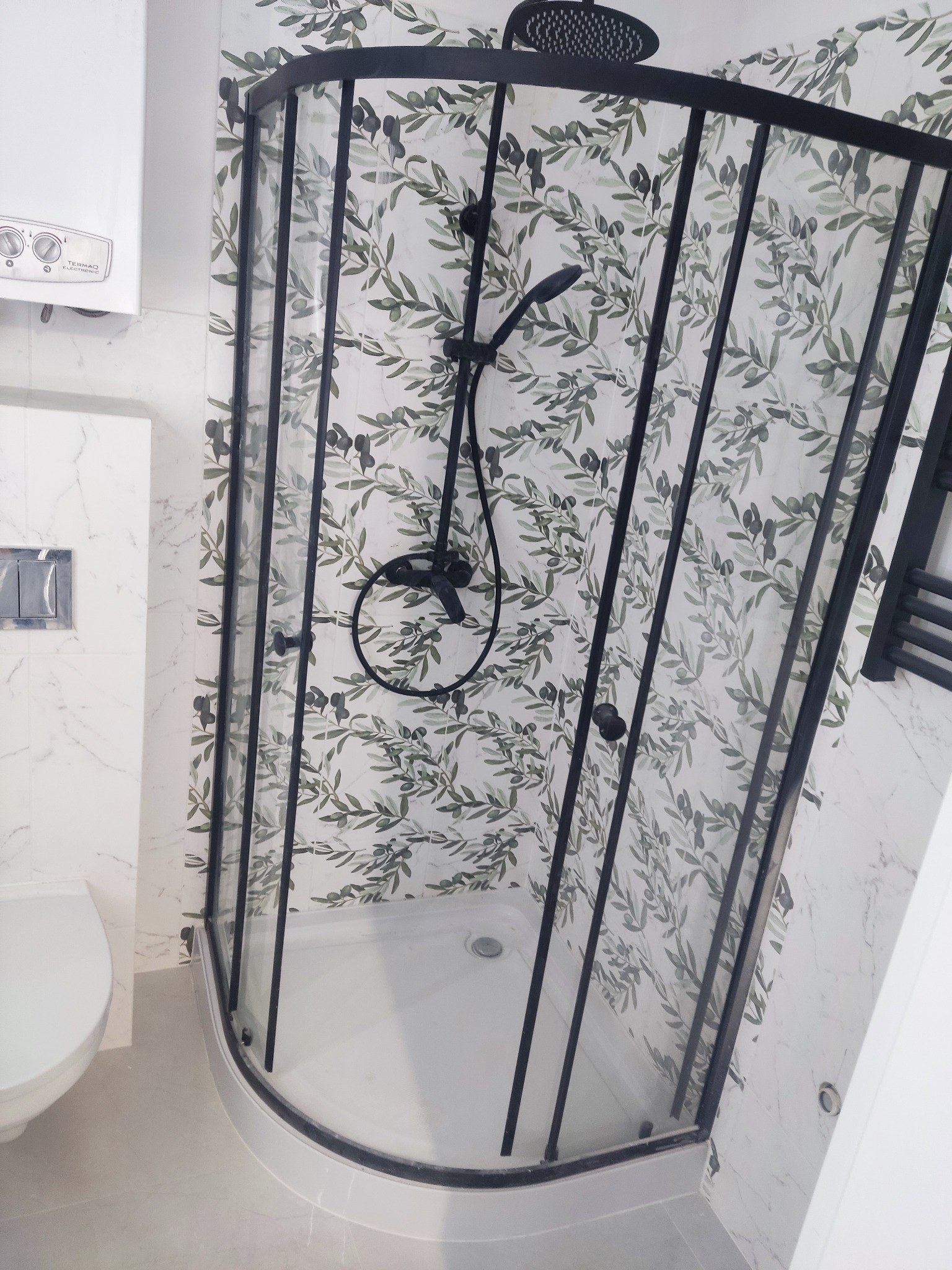 Nowoczesna kabina prysznicowa z czarną ramą i deszczownicą, na ścianie płytki z motywem liści oliwnych, widoczny bojler i fragment toalety.