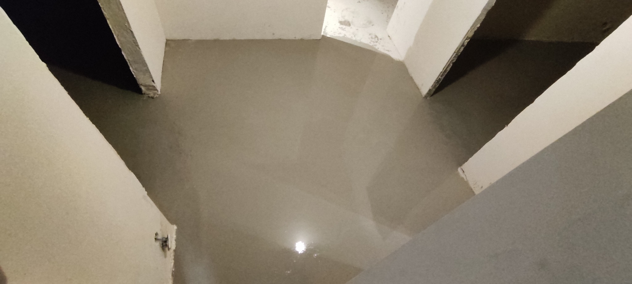 Wylana, świeża wylewka samopoziomująca w pomieszczeniu z otwartymi przejściami. Widoczne ściany w surowym stanie i odbicie światła w mokrej powierzchni.